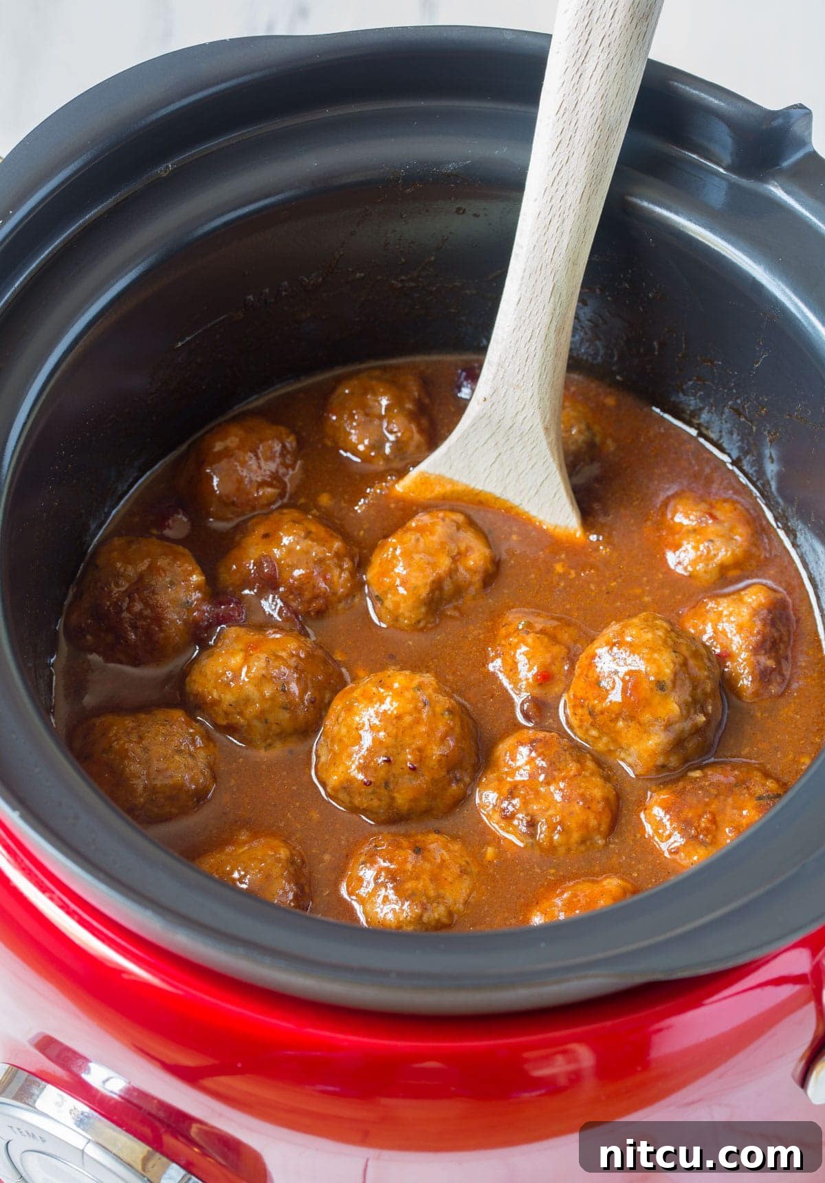 Slow cooker cocktail meatballs glistening in sauce with a wooden spoon ready to serve