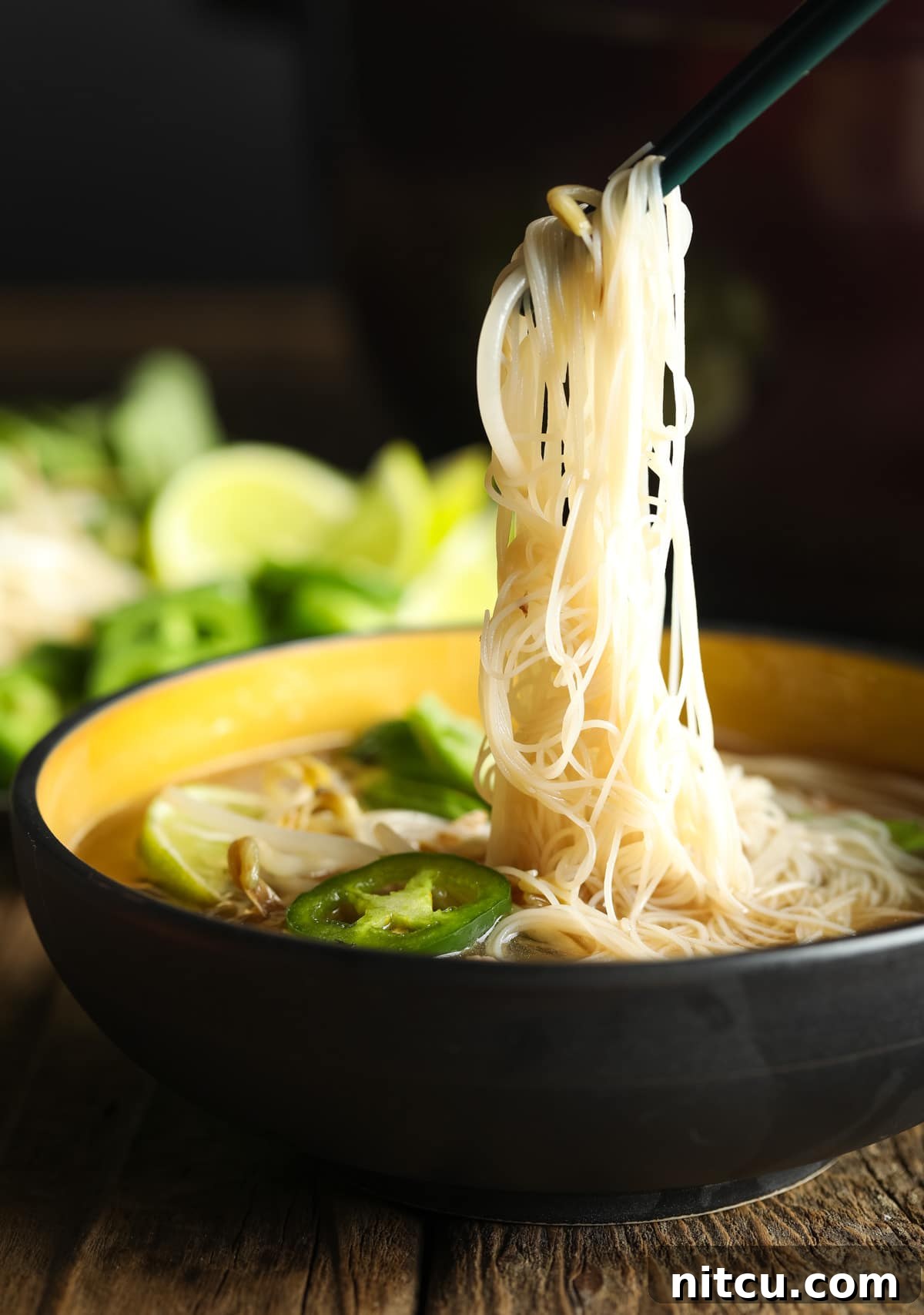 A vibrant bowl of healthy pho soup with chopsticks lifting a tangle of low-carb noodles, garnished with fresh herbs and lime.