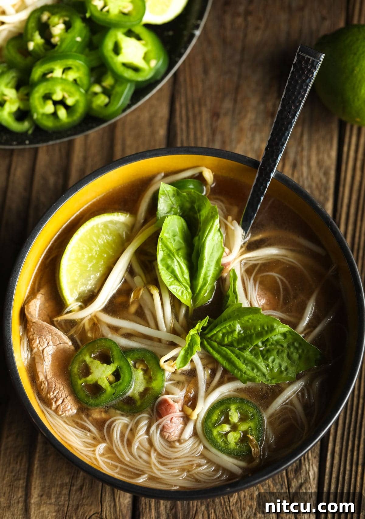 Overhead shot of a beautifully arranged healthy pho bowl, adorned with fresh basil, vibrant jalapeños, and a squeeze of lime, showcasing its rich flavors.