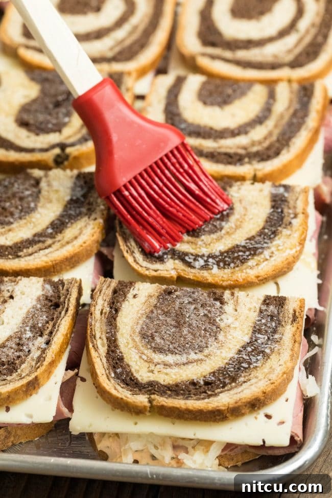 Close-up of melted butter being brushed on top of assembled Reuben sandwiches before baking.