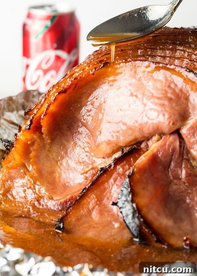 A spoon is used to drizzle the caramelized Coca-Cola glaze over slices of baked ham, highlighting its glistening texture just before serving.