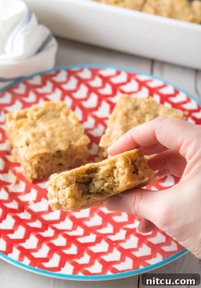 A hand holding a perfectly baked hummingbird blondie with a bite taken out, showing the moist interior.