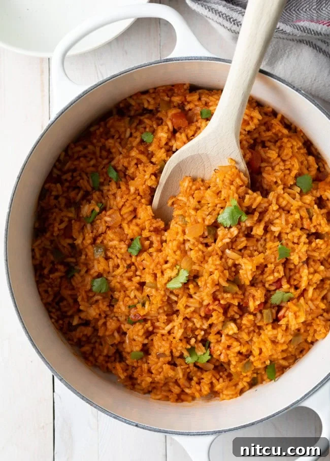 A dutch oven filled with vibrant Spanish rice, garnished with fresh cilantro, and a wooden spoon resting inside. The rice is a rich reddish-orange color, indicating a flavorful tomato base, and appears perfectly cooked and fluffy. This image showcases a delicious and inviting serving of homemade Mexican rice, ready to be enjoyed as a comforting side dish.