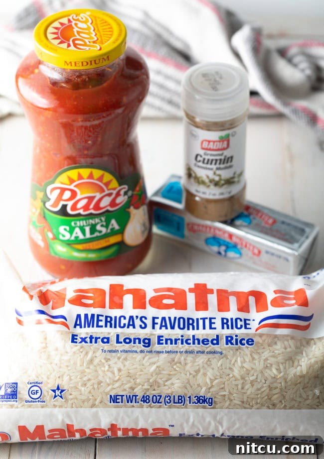 The four simple ingredients laid out for Easy Spanish Rice: a bag of long-grain white rice, a jar of chunky tomato salsa, a stick of butter, and a container of ground cumin. The vibrant colors of the salsa and the simplicity of the setup emphasize the ease of the recipe.