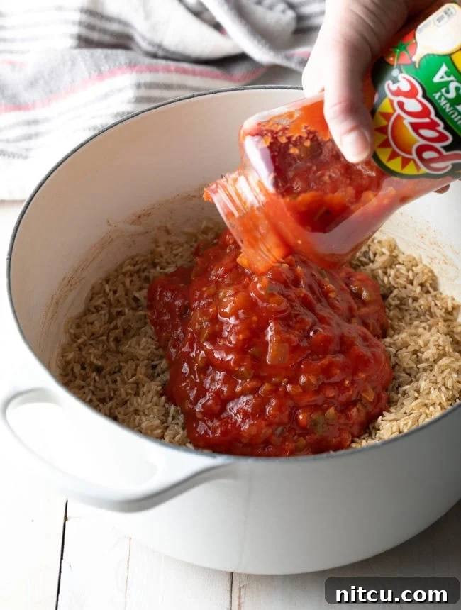 Salsa being poured from a jar into a pot with sautéed rice and butter, showing the initial mixing step for the Spanish rice recipe. The vibrant red of the salsa contrasts with the golden rice grains.