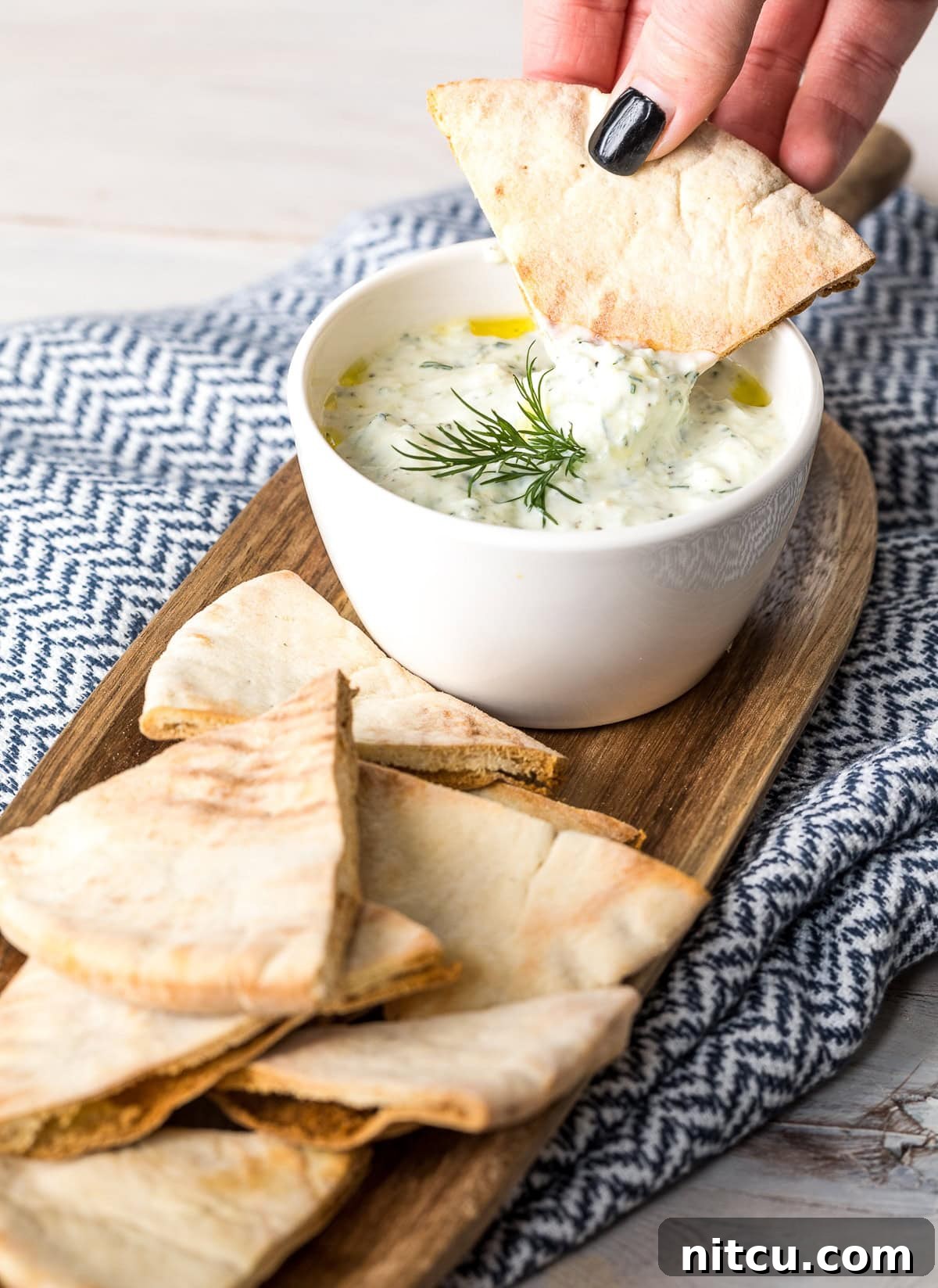 A hand dipping a piece of pita bread in tzatziki sauce that's in a white bowl.