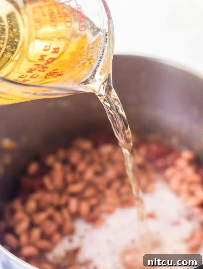 A measuring cup steadily pours a rich, amber beer into a simmering stock pot filled with chili ingredients, enhancing the depth of flavor.