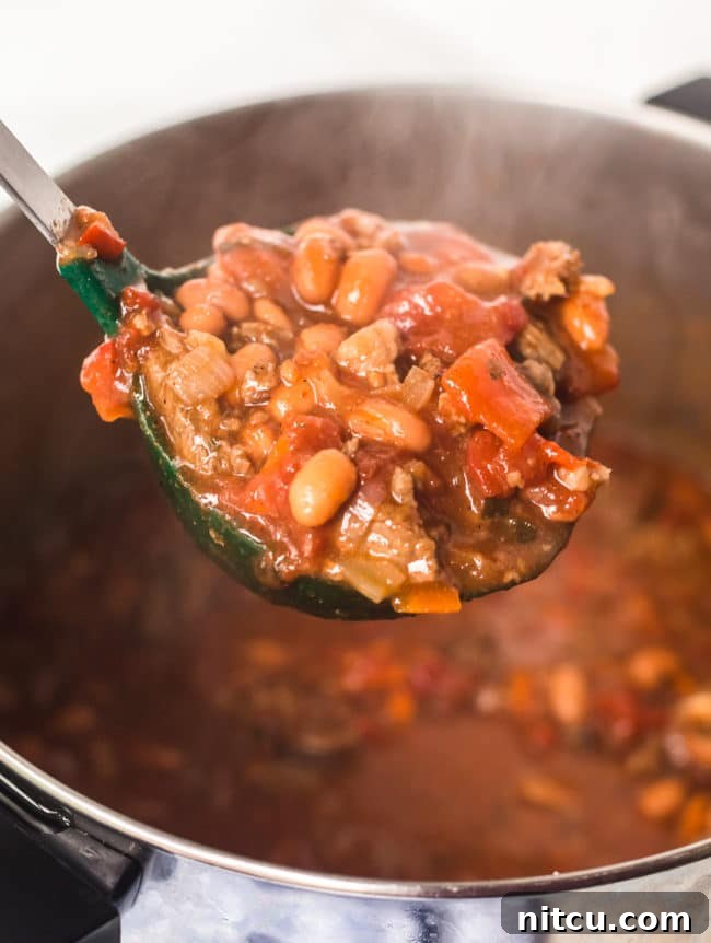 A ladle expertly scoops a generous portion of rich steak chili with beer, showcasing the hearty chunks of meat and beans, with the full pot simmering in the background.