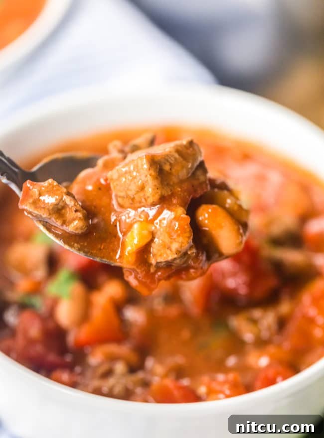 A spoon laden with rich steak chili with beer, poised above the serving bowl, highlighting the dish's irresistible texture.