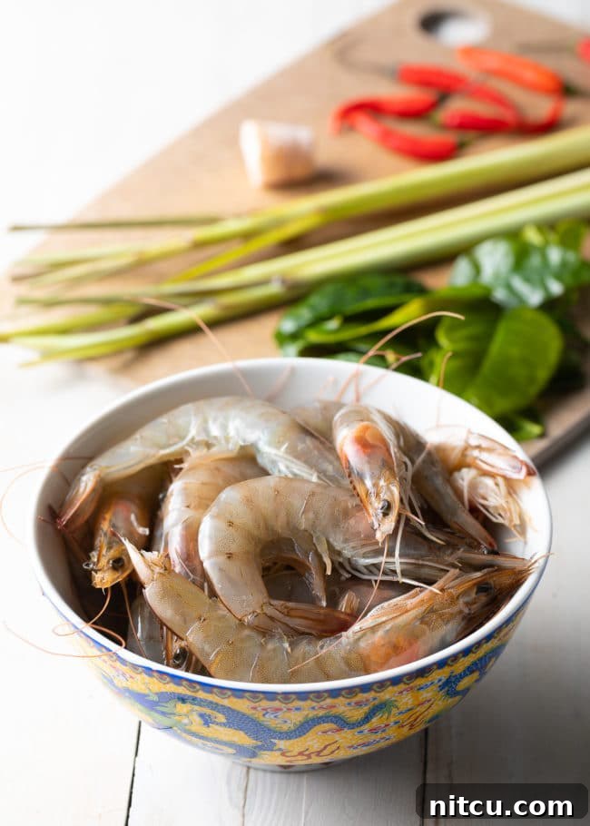 A bowl of raw shrimp arranged with fresh lemongrass, kaffir lime leaves, and other essential Tom Yum soup ingredients, ready for preparation.