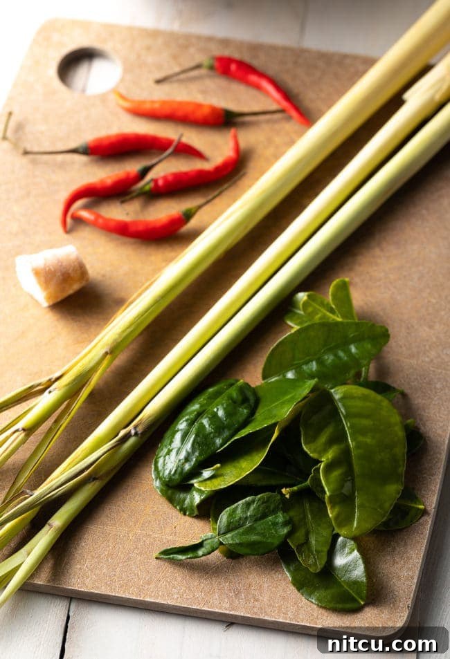 Fresh ingredients for Tom Yum soup including lemongrass, kaffir lime leaves, galangal, chiles, and onion laid out on a wooden cutting board.