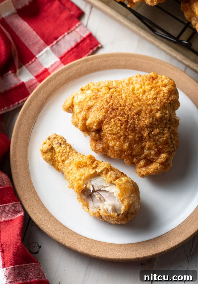 A top-down view of a breaded fried chicken thigh and drumstick on a white plate, with a bite taken from the drumstick to show the juicy interior.