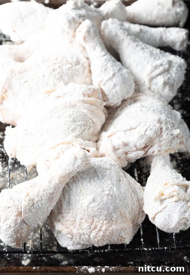 Several pieces of skin-on and bone-in chicken, freshly dredged in flour, laid out on a wire rack to rest.