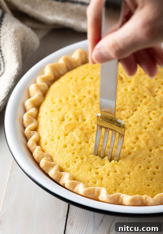 A fork pricking holes into the freshly baked sponge cake within the pie crust, preparing it to absorb the Tres Leches mixture.