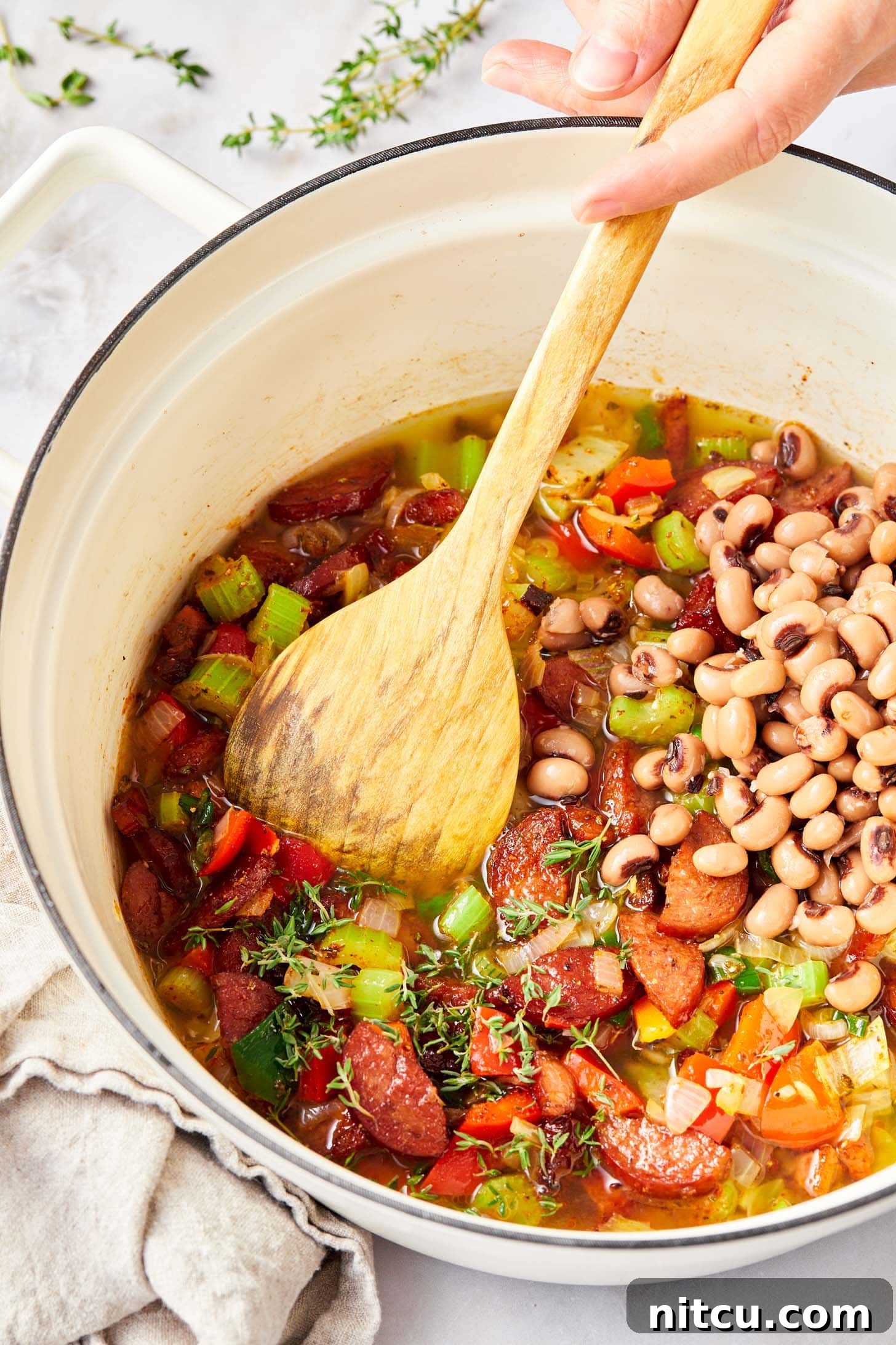 Adding black-eyed peas and chicken broth to the skillet with cooked meats and vegetables, ready for simmering.