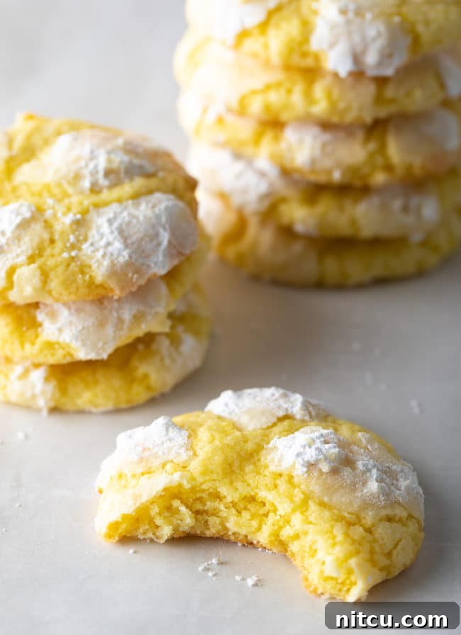 A delicious Lemon Crinkle Cookie with a bite taken out, revealing its soft interior, with more cookies stacked beautifully in the background.