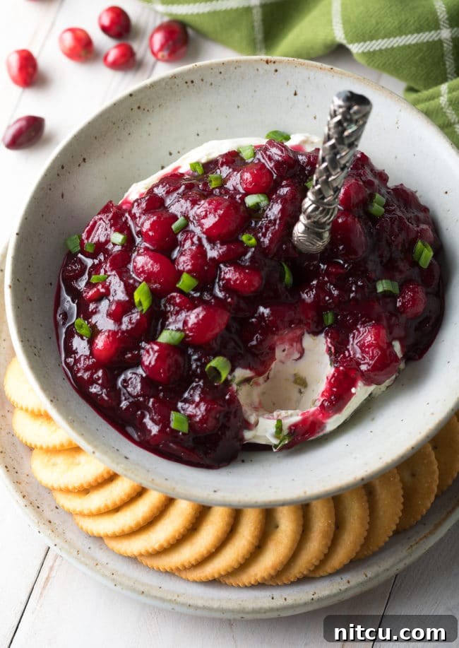 Close-up of Cranberry Jalapeno Cream Cheese Dip in a serving bowl, showcasing its layered texture with a serving spoon poised to scoop, emphasizing its creamy and flavorful appeal.