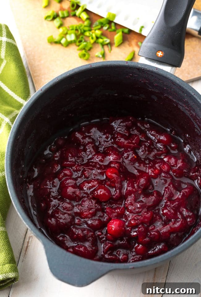 Pomegranate cranberry sauce simmering in a saucepot, depicting the rich color and thickening consistency of the topping.