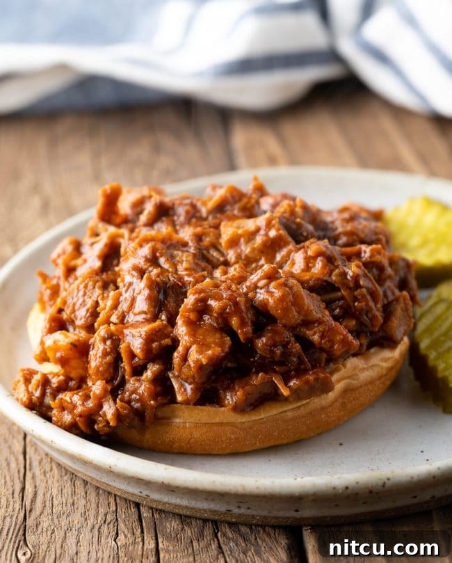An open-face chopped beef brisket sandwich on a soft bun, garnished with pickles, ready to be assembled.