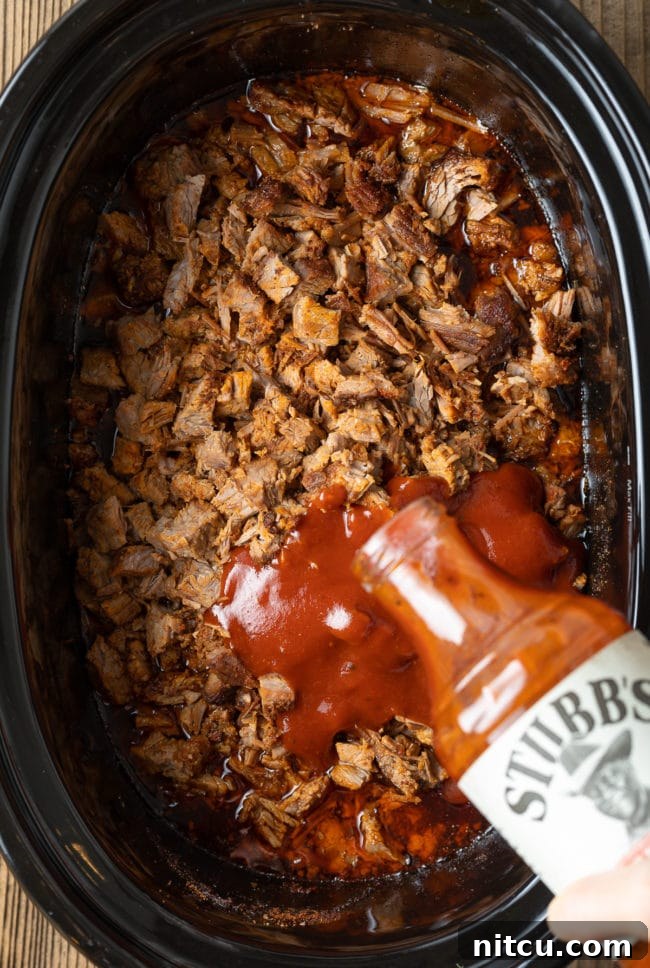 A generous amount of spicy BBQ sauce being poured over finely chopped beef in a slow cooker.
