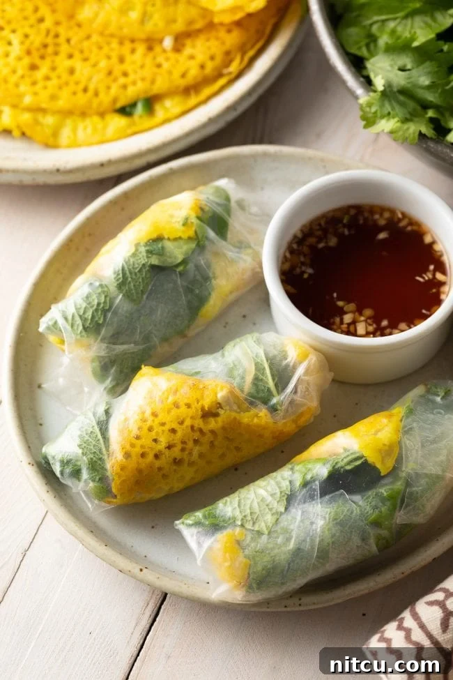 A vibrant plate featuring three golden Bánh Xèo triangles, beautifully wrapped in delicate rice paper and fresh herbs, with a small bowl of rich brown Nuoc Cham dipping sauce on the side.