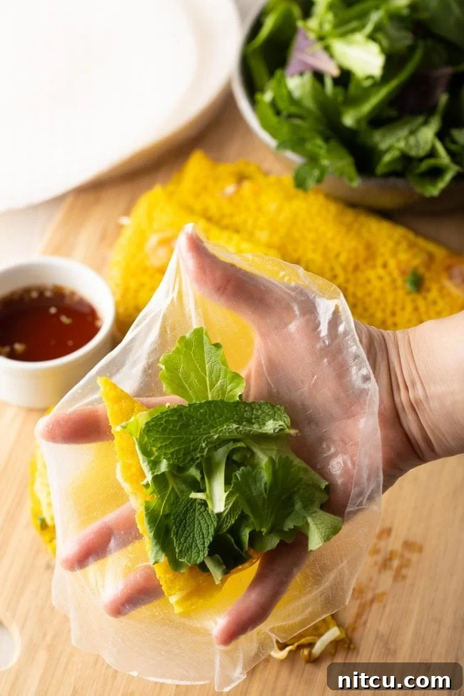 A hand layering fresh mint leaves onto a Bánh Xèo triangle placed on a rice paper sheet, preparing it for wrapping.