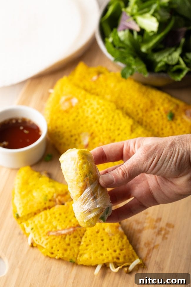 A hand proudly presents a perfectly rolled Bánh Xèo spring roll, showcasing the crisp crepe and fresh herbs encased in transparent rice paper, ready to be dipped.
