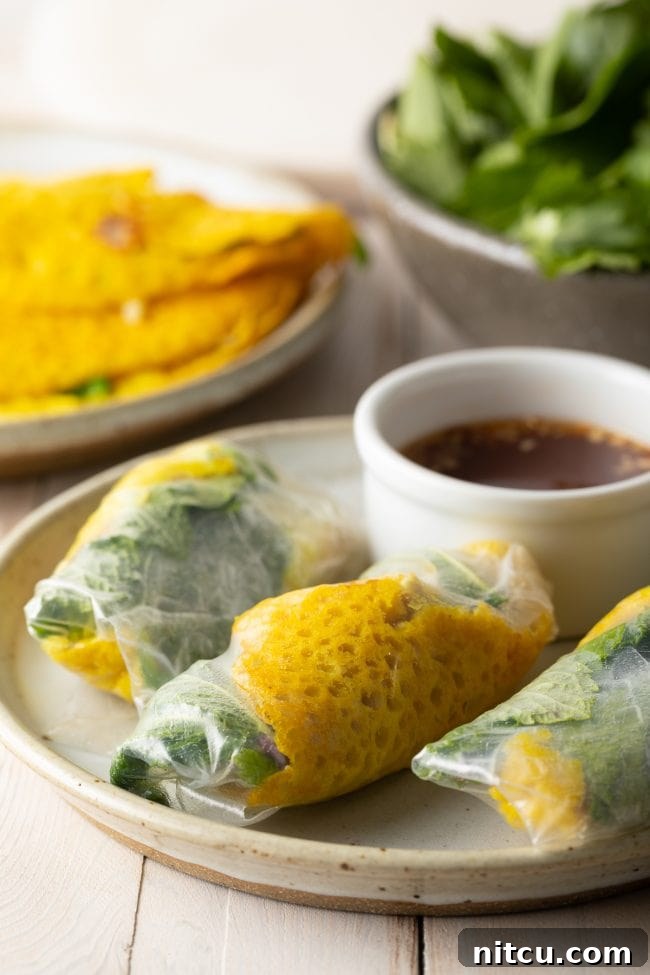 A dish featuring three beautifully arranged Bánh Xèo, each wrapped in rice paper and ready to eat, with a small ramekin of dipping sauce placed beside them.
