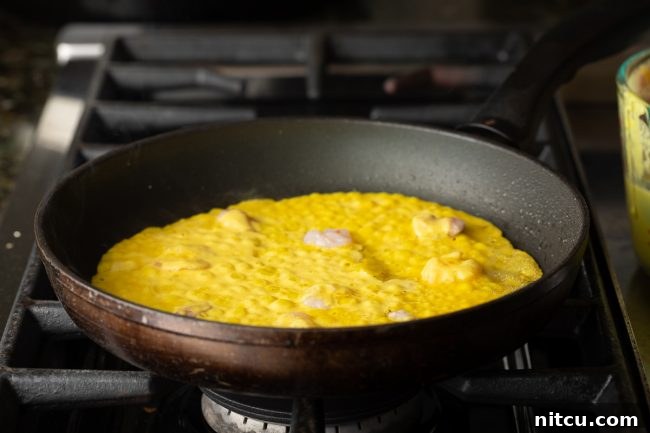 A ladle pours thin, golden-yellow Bánh Xèo batter over sizzling shrimp and pork in a hot skillet, coating the bottom evenly.