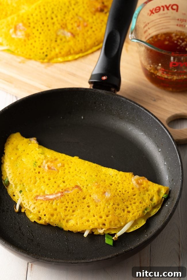 A perfectly folded, golden-brown Vietnamese crepe (Bánh Xèo) sits in a hot skillet, its edges crisp and inviting, showcasing the delicious filling within.