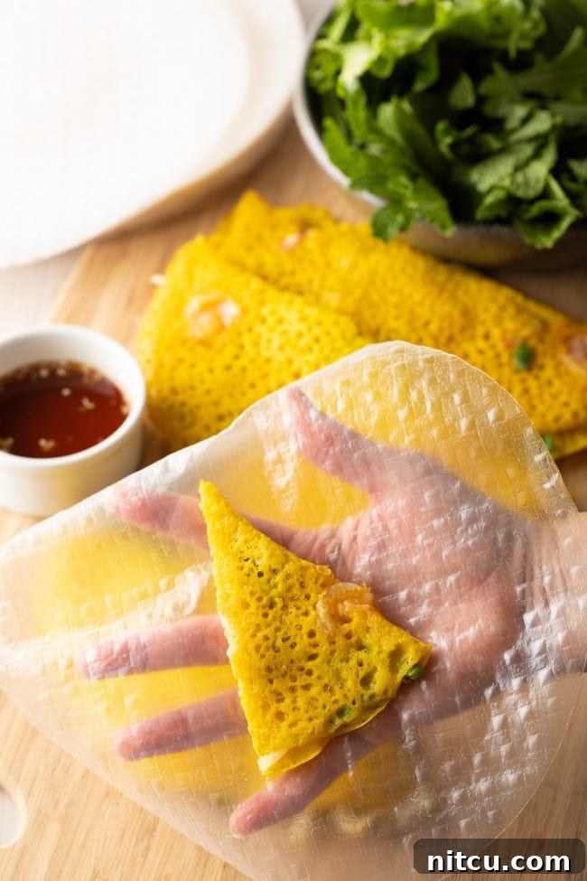 A hand holds a large, translucent rice paper sheet with a triangular piece of golden Bánh Xèo centered on it, ready for wrapping.