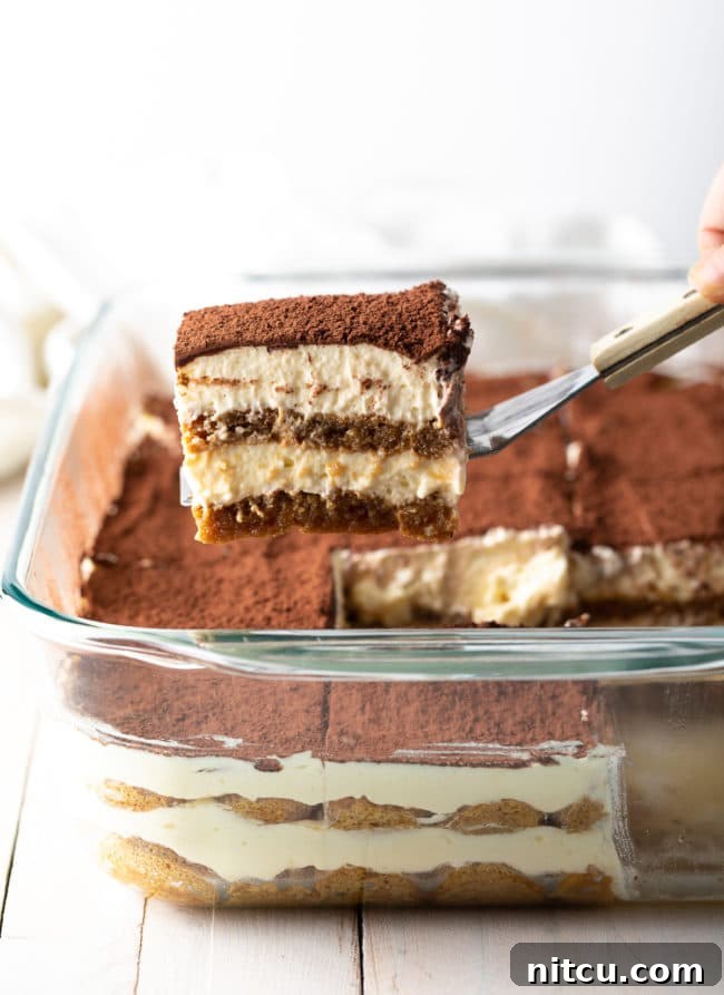 A perfectly sliced piece of classic Tiramisu held on a spatula, with a larger tray of Tiramisu visible in the background, showcasing its rich layers of cream and coffee-soaked biscuits.