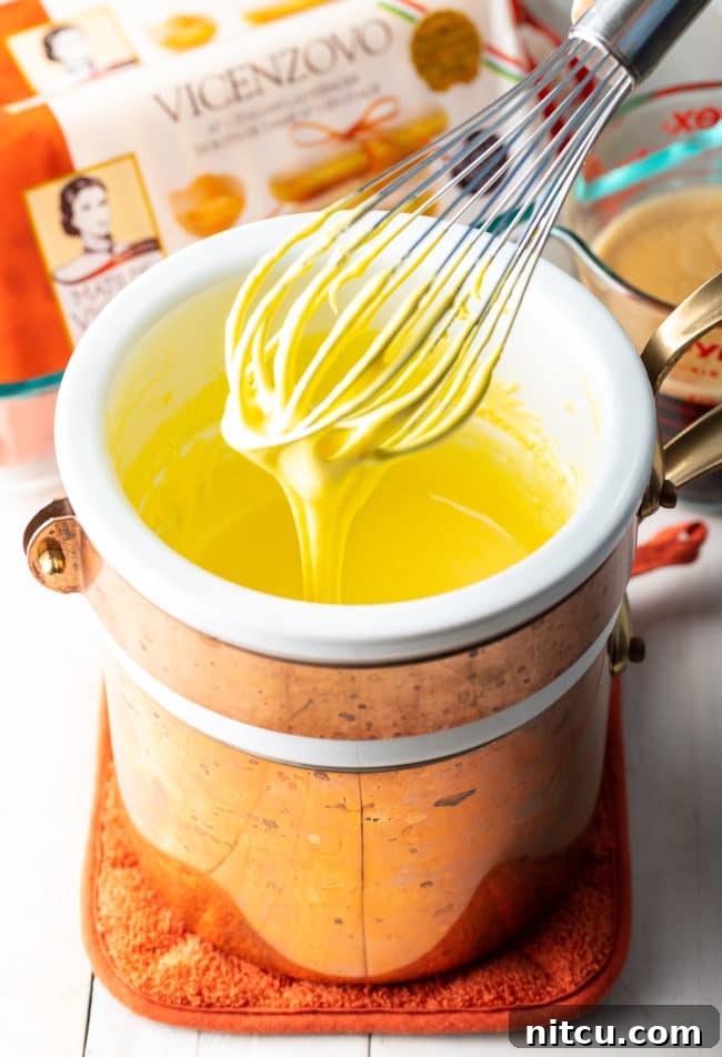A whisk vigorously mixing egg yolks and sugar in a bowl over a double boiler, creating the silky smooth sabayon for the tiramisu cream.