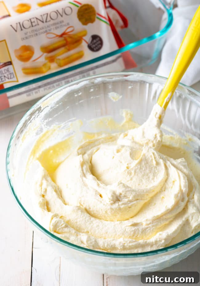 A rubber spatula gently folding mascarpone and whipped heavy cream together in a glass bowl, ensuring a light and airy texture for the tiramisu filling.