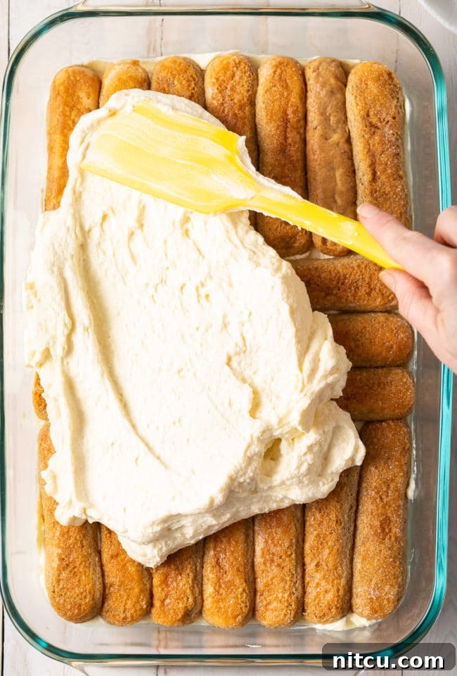 A spatula smoothly spreading a generous layer of creamy mascarpone mixture over the coffee-soaked ladyfingers in the baking dish, creating the iconic layers.