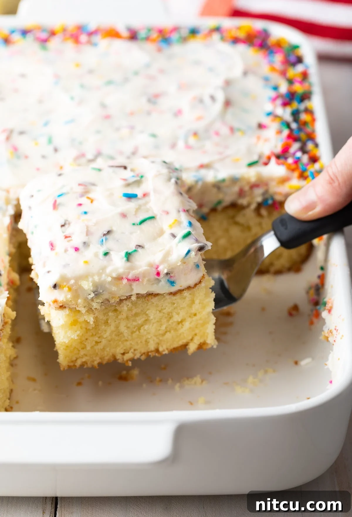 A hand holding a spatula, lifting a perfect piece of vanilla cake out of the baking dish.