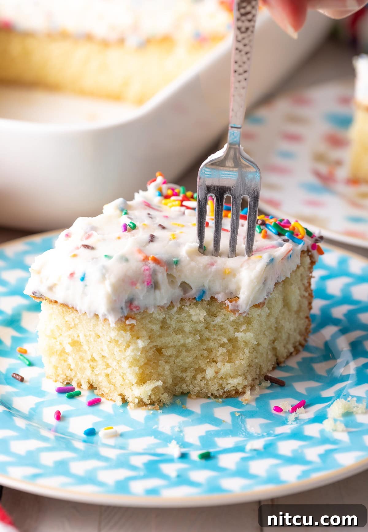 A slice of vanilla cake on a plate with a fork poised to take a bite.