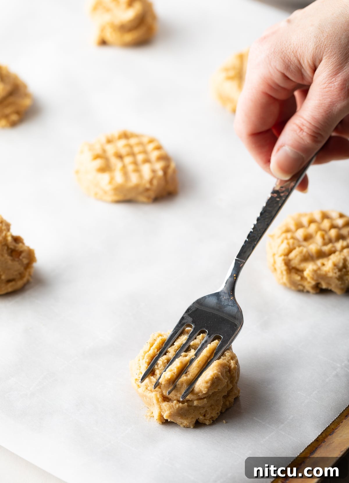The classic criss-cross pattern being pressed onto peanut butter cookie dough balls with a fork on a baking sheet, ready for the oven.
