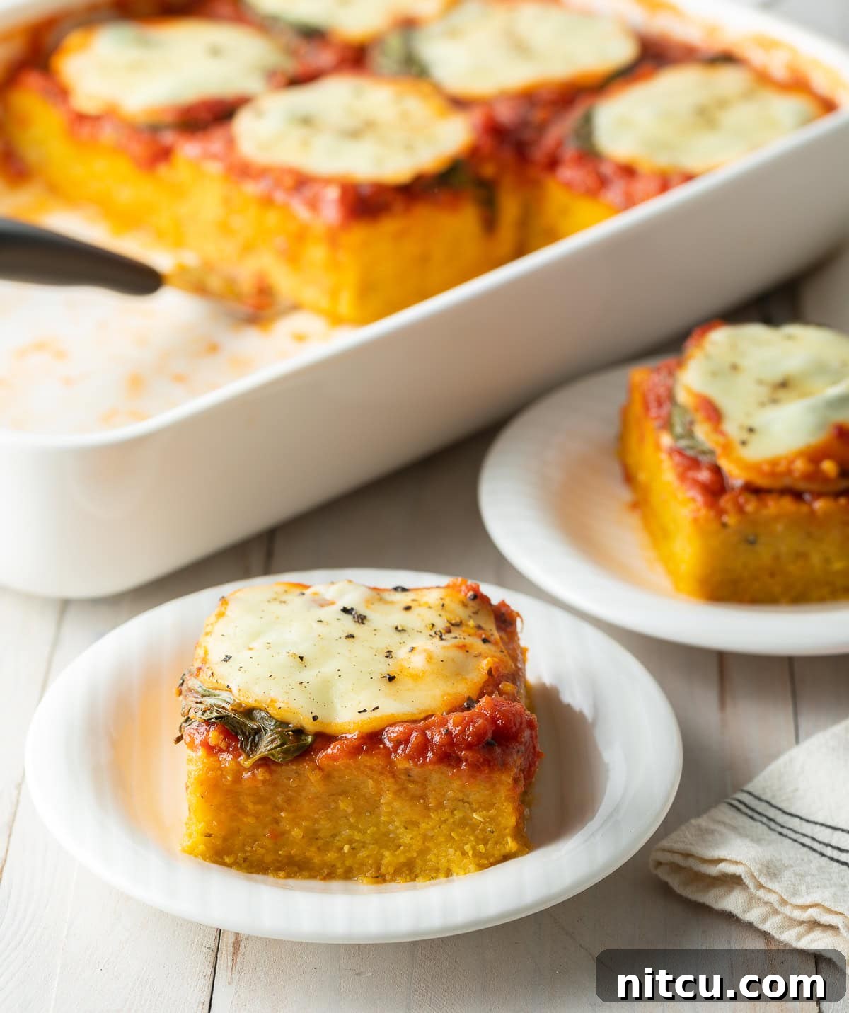 A baking dish filled with golden-baked polenta, topped with rich marinara sauce, fresh green basil leaves, and thick slices of melted mozzarella cheese. A perfect square of Margherita baked polenta sits ready to serve on a white plate in the foreground.