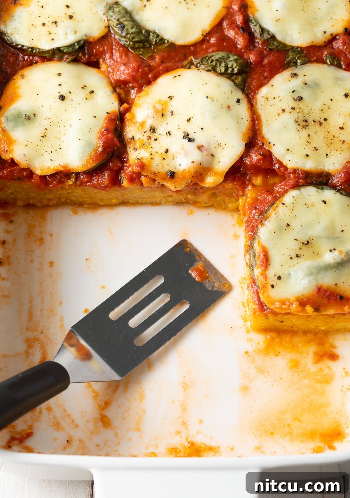 A top-down view of a baking dish filled with freshly baked polenta, showcasing a removed slice, revealing its creamy, layered interior and golden crust.