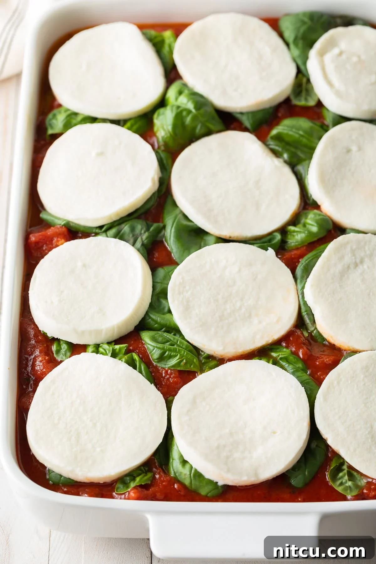 A top-down view of a baking dish, showcasing the firm baked polenta base topped with vibrant uncooked marinara sauce, fresh basil leaves, and generous slices of fresh mozzarella, ready for the final bake.