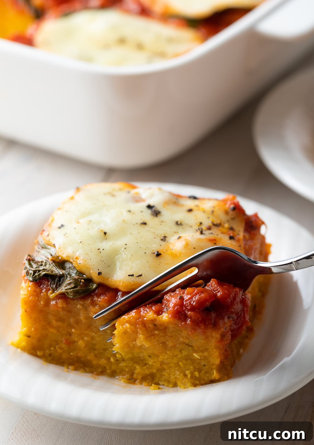 A fork lifts a perfectly cut chunk of cheesy baked polenta from a plate, showing its inviting layers.