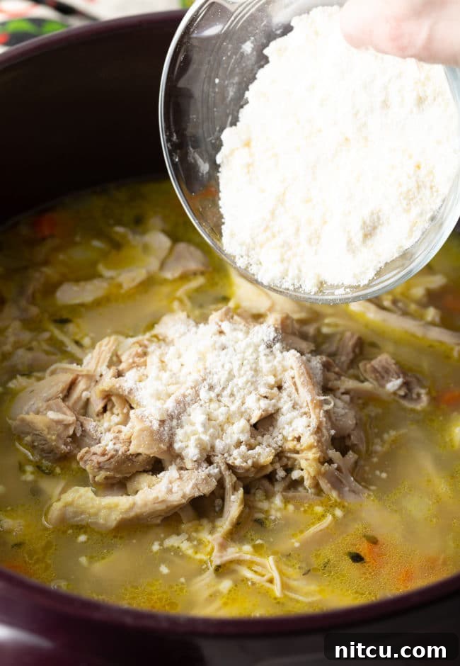 A hand pouring a mixture of cornstarch and grated Parmesan cheese into a pot of simmering chicken and rice soup.
