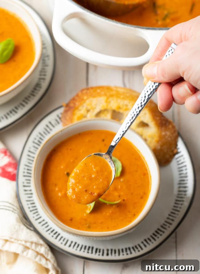 A bowl of creamy roasted tomato soup with a hand holding a spoonful of the soup above the bowl, ready to be tasted.