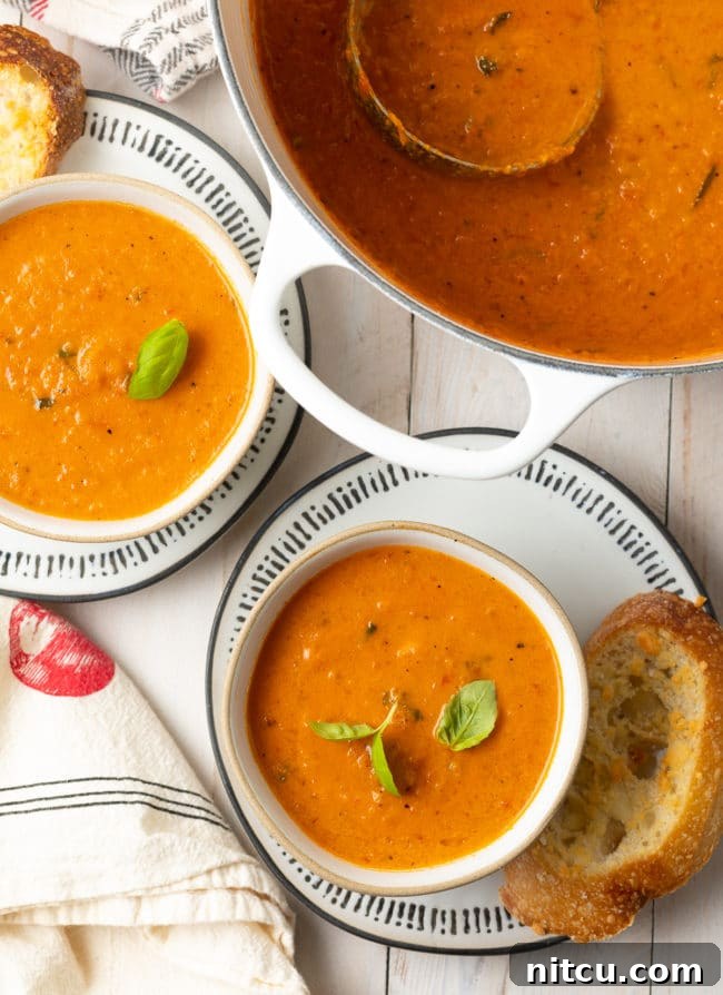 Two elegantly presented bowls of creamy roasted tomato soup, with a large pot of the soup simmering gently in the background.