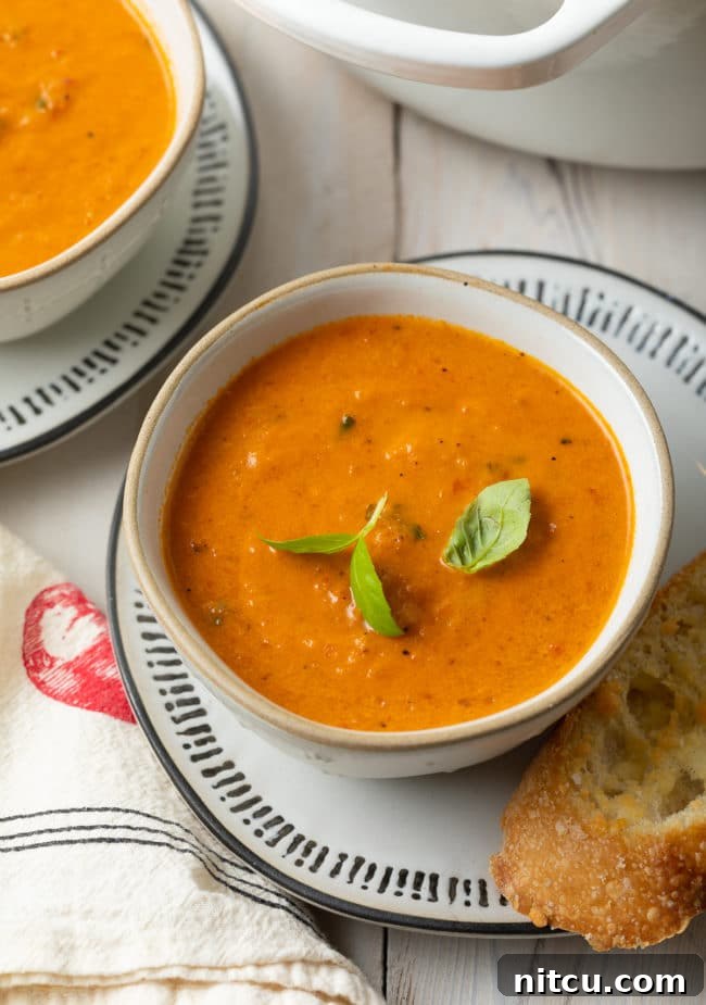 A bowl of creamy roasted tomato soup garnished with fresh basil sprigs, accompanied by a slice of crusty bread on the side.