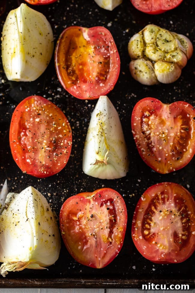 Fresh plum tomatoes, a sweet onion, and a head of garlic arranged on a baking sheet, ready for roasting.