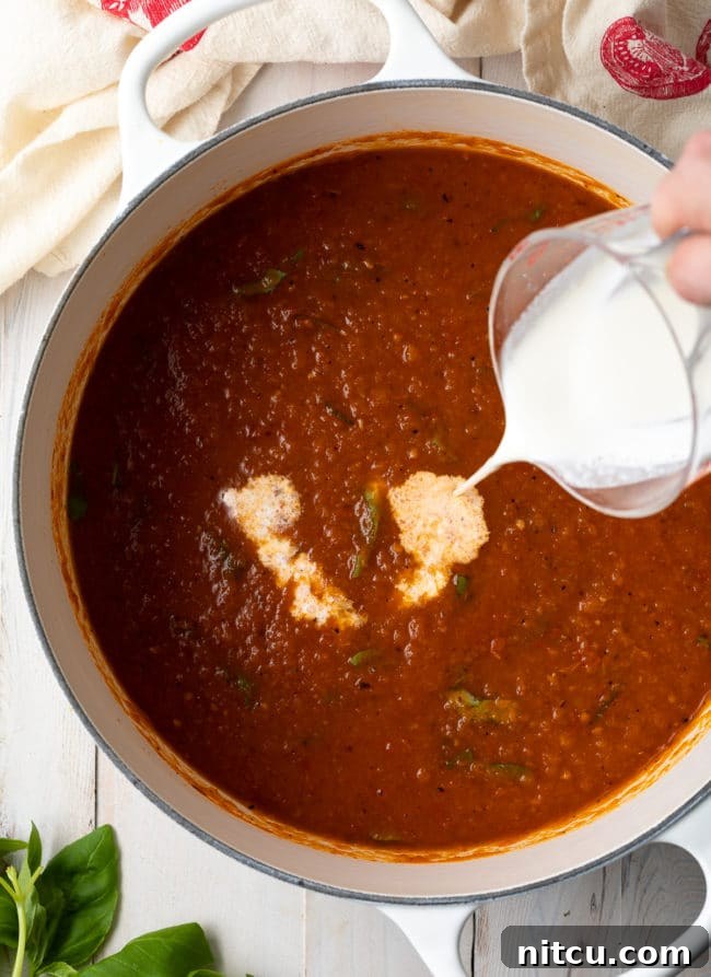 A hand gently pouring heavy cream into a simmering pot of rich tomato soup, adding to its creamy texture.