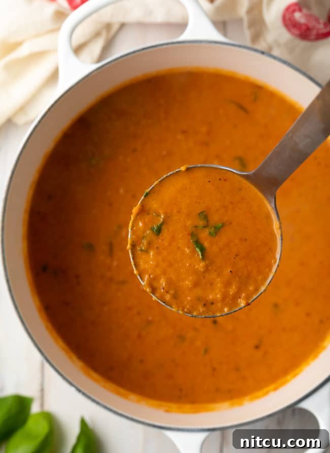 A ladle filled with creamy roasted tomato soup, held above the pot, showcasing its smooth and inviting texture.