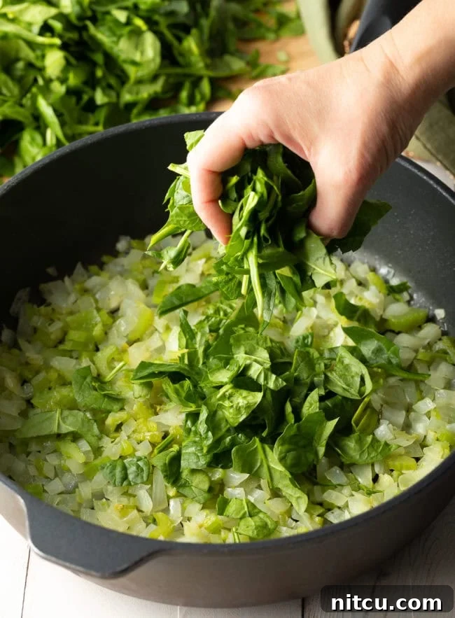 A large pot filled with sautéed onions, celery, and chopped fresh spinach, slowly wilting down to create the flavorful base for the pie filling.